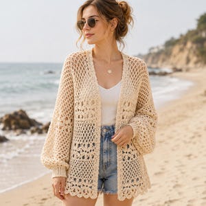 May include: A cream-colored open-front crochet cardigan featuring a repeating floral motif pattern, long balloon sleeves with ribbed cuffs, and a scalloped hemline. The garment is worn over a white tank top and light blue denim shorts.