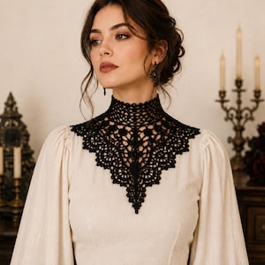 May include: A cream-coloured long-sleeved blouse featuring a high-neck black lace collar. The intricate black lace overlay covers the upper chest and neck area, showcasing a detailed floral pattern with a pointed hemline at the centre.