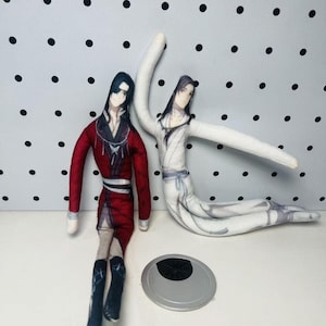 May include: Two handmade fabric dolls. One doll is dressed in a red and black outfit, the other in a white outfit. The dolls are on a white surface with a silver object in front of them. The background is white with black polka dots.
