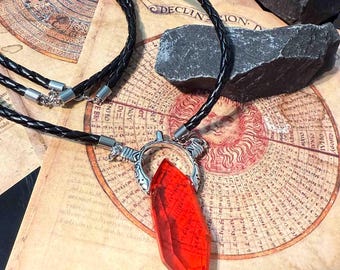 May include: A pendant necklace featuring a large, faceted red crystal. The crystal is suspended from a silver-toned metal setting and hangs from a black braided leather cord. The background includes a vintage map and dark gray stones.