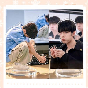 May include: Two acrylic standees featuring images of a person in different poses. One shows a person in a blue shirt and khaki pants, the other shows a person in a black shirt holding a cup. Both standees have a clear circular base.