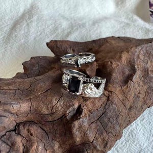 May include: Two silver rings with black gemstones are displayed on a piece of dark wood. The larger ring features a rectangular black stone, while the smaller ring has a smaller, rectangular black stone. Both rings have intricate detailing.
