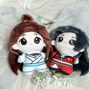 May include: Two plush dolls with large eyes and detailed facial features. One doll has brown hair and a white outfit with blue accents. The other doll has gray hair and a red and white outfit. Both dolls are on a white background.
