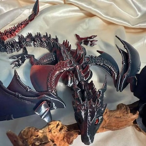 May include: A detailed, 3D-printed dragon sculpture in black and red. The dragon features intricate scales, wings, and a menacing head with red eyes. The sculpture is posed on a piece of driftwood, with a satin background.