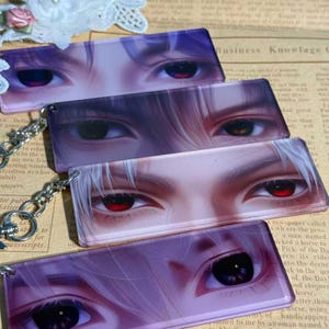 May include: Four rectangular keychains featuring anime-style eyes with varying colors. The eyes have detailed shading and highlights, with colors including red, brown, and black. Each keychain has a silver-colored chain attached.