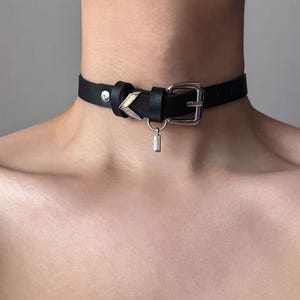 Puede incluir: Collar gargantilla de cuero negro con hebilla plateada y un pequeño dije rectangular. La gargantilla presenta un acento decorativo en forma de cheurón. El collar está diseñado para usarse alrededor del cuello.