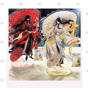 Puede incluir: Dos figuras de acrílico con personajes estilo anime. Un personaje viste túnicas rojas con un paraguas rojo, el otro viste túnicas blancas y amarillas con un paraguas blanco. Ambos tienen cabello largo y oscuro y bases decorativas.