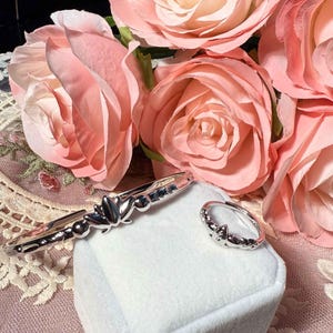 Könnte beinhalten: Ein silbernes Armband- und Ring-Set mit floralem Design, präsentiert auf einer weißen Oberfläche. Der Schmuck ist vor einem Hintergrund aus rosa Rosen platziert. Das Armband hat ein zentrales Lotusblüten-Design.