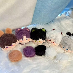 Puede incluir: Una colección de llaveros de peluche con caras de personajes de dibujos animados con varios colores de pelo, incluyendo marrón, morado, negro, blanco y gris. Cada llavero tiene un pompón a juego. Los llaveros se muestran sobre una superficie blanca y esponjosa.
