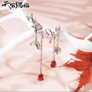 May include: Silver earrings with a floral and leaf design. Each earring has a chain with a red teardrop-shaped gem. The earrings have a delicate, elegant appearance, suitable for special occasions.