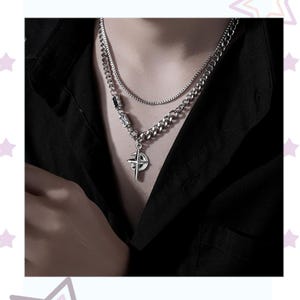 May include: A silver-toned necklace set featuring a curb chain and a thinner box chain. The pendant is a compass design. The necklace is worn against a black shirt.