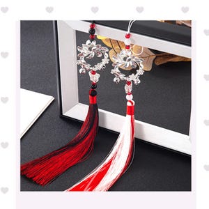 May include: Two decorative car charms with silver floral designs and long tassels. One tassel is red and black, the other is red and white. The charms hang from a white string and have red and black beads.