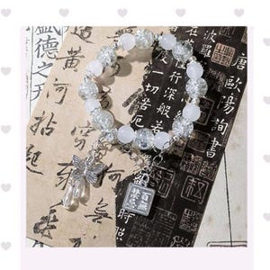 Puede incluir: Una pulsera delicada con cuentas blancas y transparentes, acentuada con un dije de mariposa plateada y un colgante rectangular con caracteres asiáticos. La pulsera se exhibe sobre un fondo de papel envejecido con caligrafía asiática.