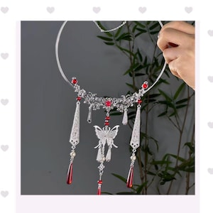 Puede incluir: Un collar plateado con intrincados diseños florales y de mariposas. El collar presenta detalles de piedras preciosas rojas y colgantes en forma de lágrima. El diseño incluye una delicada cadena y es sostenido por una mano.