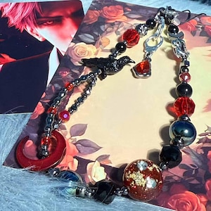 Könnte beinhalten: Ein Perlenarmband mit einem Rabenanhänger, roten und schwarzen Perlen und einem roten Halbmond. Das Armband umfasst eine Vielzahl von Perlenformen und -größen mit silberfarbenen Metallakzenten. Der Hintergrund zeigt ein Blumendesign.