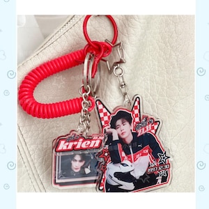May include: A keychain with two clear acrylic charms featuring images of a person. One charm has the word "krien" and the other has the date "1997.12.30". The keychain has a red wrist strap and silver-coloured metal rings.