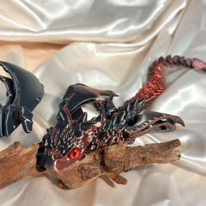 Op de afbeelding: Een gedetailleerd drakenbeeldje in zwart en rood, met een rood oog, rustend op drijfhout. De draak heeft ingewikkelde vleugel- en lichaamsdetails, met een rode gradiënt op zijn staart. Het beeldje staat tegen een witte satijnen achtergrond.
