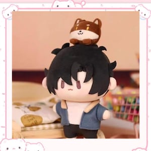 Peut inclure: Une poupée en peluche avec des cheveux noirs, une veste bleue et un petit chien en peluche marron sur le dessus. La poupée porte une chemise blanche et un pantalon noir. La poupée est sur un lit avec un oreiller avec le texte "Since 1988".