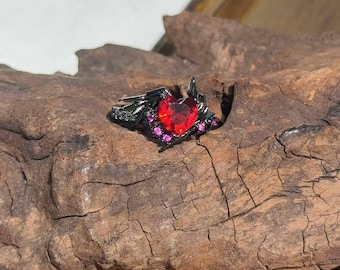 May include: A black ring with a heart-shaped red gemstone at the center, flanked by smaller pink gems. The ring has a detailed, gothic-inspired design, set against a textured wooden surface.