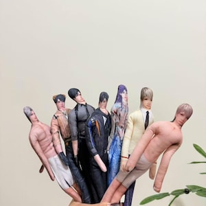 Könnte beinhalten: Eine Sammlung handgefertigter Stoffpuppen, jede mit einem einzigartigen Outfit und Frisur. Die Puppen variieren in Hautton und Kleidungsstil, darunter ein Anzug, eine Jacke und eine Puppe ohne Hemd. Die Puppen werden zusammengehalten.