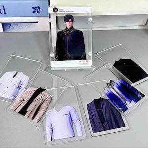 Puede incluir: Expositores transparentes de acr&iacute;lico que muestran im&aacute;genes de ropa y una persona. La ropa incluye una bata de laboratorio blanca, una gabardina beige, una camiseta de cuello en V de color p&uacute;rpura claro y un blazer azul oscuro. La persona lleva un atuendo negro.