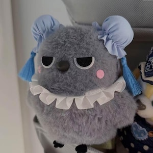 Puede incluir: Un peluche gris con una expresión de mal humor, ojos negros y mejillas rosadas. Tiene lazos azules en la cabeza con borlas a juego y un cuello blanco con volantes. El juguete está hecho de un material suave y esponjoso.