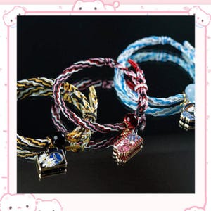 Puede incluir: Primer plano de varias pulseras tejidas en varios colores, incluyendo rojo, blanco, azul y amarillo. Cada pulsera presenta un dije decorativo, como una pequeña caja con detalles florales o un edificio en miniatura con acentos rosas. Las pulseras se exhiben sobre un fondo oscuro.
