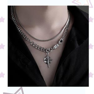 May include: A silver-toned layered necklace with a compass pendant. The necklace features a combination of chain styles, including a curb chain and a thinner, more delicate chain. The pendant has a compass design with a cross and star details. The necklace is worn against a black shirt.