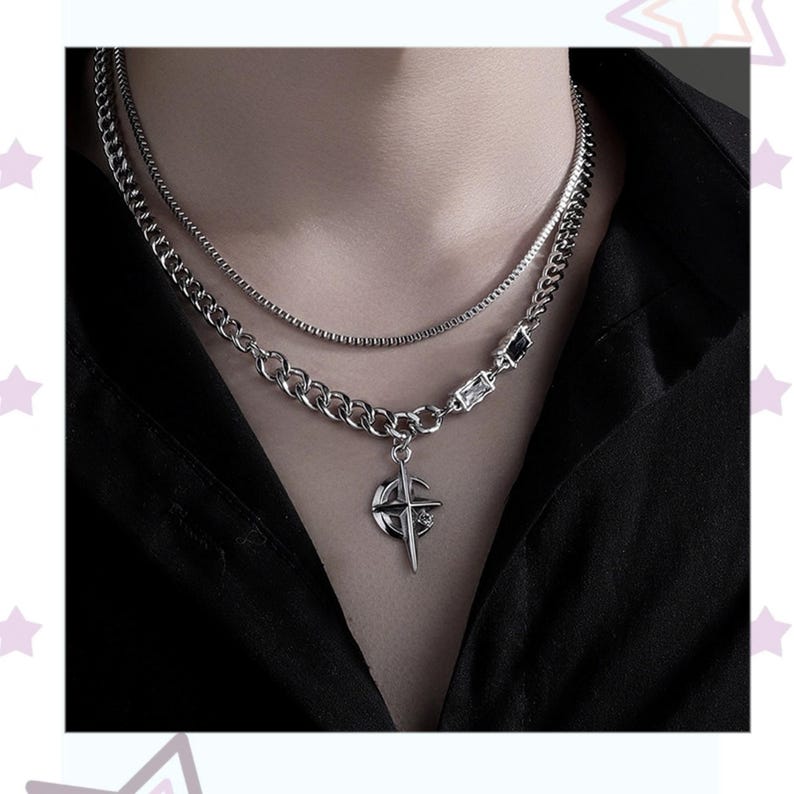 May include: A silver-toned layered necklace with a compass pendant. The necklace features a combination of chain styles, including a curb chain and a thinner, more delicate chain. The pendant has a compass design with a cross and star details. The necklace is worn against a black shirt.