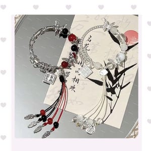 May include: Two silver bracelets with butterfly details and dangling tassels. One bracelet has red and black beads, the other clear and white. Each bracelet features a small square charm with Chinese characters.