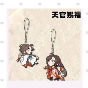 May include: Two anime-style character charms with attached lanyards. One character is in white and blue clothing, the other in red and orange. The text "Heaven Official's Blessing" is above the charms.