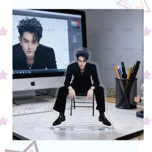 May include: A clear acrylic stand-up figure featuring a man in a black suit seated on a small chair. The figure is in front of a computer screen displaying a similar image. Pens and pencils are in a holder to the right.