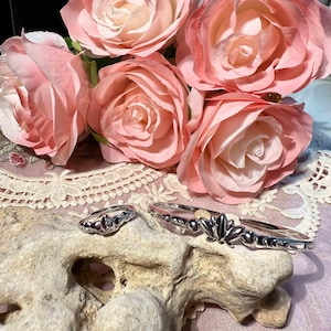 Könnte beinhalten: Ein silbernes Ring- und Armband-Set mit floralem Design, präsentiert auf einem strukturierten Stein. Zarte rosa Rosen sind im Hintergrund, mit einer Spitzendecke darunter. Der Schmuck hat eine polierte Oberfläche.