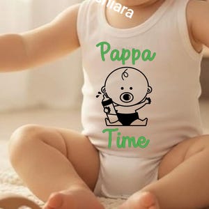 Puede incluir: Un body blanco sin mangas para bebé con el texto verde Pappa Time y un dibujo lineal negro de un bebé sosteniendo un biberón. El texto @etlabchiara está impreso en diagonal sobre la parte superior del pecho de la prenda.