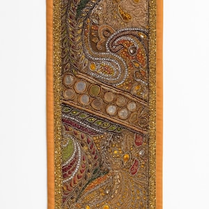 May include: A rectangular, ornate textile with a detailed paisley design. The central portion features a brown background with intricate patterns in gold, orange, green, and red. The border is a solid orange with gold trim, creating a rich, decorative piece.