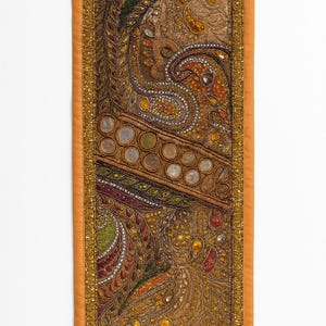 May include: A rectangular, ornate textile with a detailed paisley design. The central portion features a brown background with intricate patterns in gold, orange, green, and red. The border is a solid orange with gold trim, creating a rich, decorative piece.