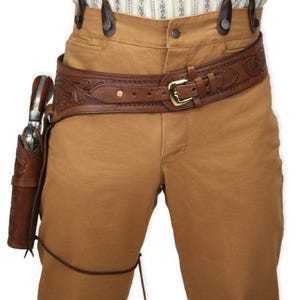 Könnte beinhalten: Ein brauner Leder-Gürtel mit Holster, mit aufwendigen floralen Verzierungen. Der Gürtel hat eine goldfarbene Schnalle und wird mit einer beigefarbenen Hose und Hosenträgern getragen. Ein Revolver befindet sich im Holster.