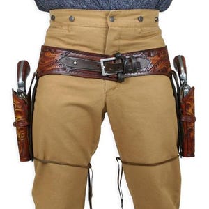 Op de afbeelding: Beige broek met een bruine leren riem en twee holsters, elk met een revolver. De riem heeft een reliëfpatroon en een zilveren gesp. De holsters zijn van bruin leer.