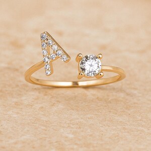 Peut inclure: Une bague dorée avec un design à anneau fendu. Un côté présente la lettre "A" incrustée de petits pierres précieuses claires. L'autre côté contient une grosse pierre précieuse claire et ronde. La bague est posée sur un fond neutre.