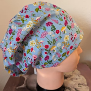 May include: A light blue fabric hat with a floral pattern featuring red, pink, yellow, and white flowers, along with green and yellow mushrooms. The hat is designed to be worn on the head.