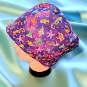 May include: A purple scrub cap featuring a colorful pattern of sea creatures including seahorses, starfish, sea turtles, and various fish. The fabric has a vibrant, multi-colored print set against a deep purple background with a fitted design.