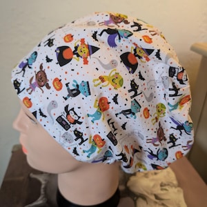 May include: White Halloween scrub cap with a repeating pattern of cartoon ghosts, bats, pumpkins, and other spooky characters. The cap is designed to fit comfortably and securely, ideal for healthcare professionals or anyone wanting a festive touch.