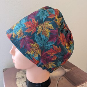 Pode incluir: Uma touca cirúrgica azul-petróleo com um padrão de folhas de outono coloridas em tons de vermelho, amarelo e roxo. A touca é exibida em uma cabeça de manequim. As folhas são detalhadas e criam um tema sazonal.