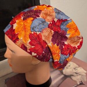 May include: A colorful, patterned hat featuring a vibrant autumn leaf design. The hat showcases a mix of red, orange, blue, and purple leaves on a cream background. This accessory is designed to be worn on the head.