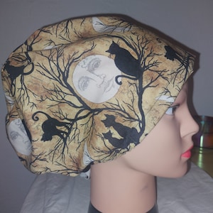 May include: A beige and brown patterned cap featuring black cat silhouettes, bare tree branches, and a full moon with a face. The cap is worn by a mannequin head. The cap is likely for medical professionals or those in the food industry.
