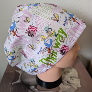 May include: A white and pink patterned scrub cap featuring illustrations of characters from Alice in Wonderland. The cap includes the text "That Way" and "Go back!" in green and blue. The design includes the Mad Hatter, Alice, and the Cheshire Cat.