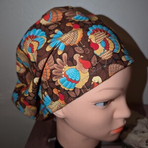 Pode incluir: Uma touca cirúrgica castanha com um padrão repetido de perus coloridos. Os perus têm detalhes azuis, vermelhos, laranja e amarelos. A touca foi concebida para cobrir a cabeça e tem um padrão por toda a parte.