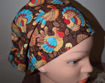 Könnte beinhalten: Eine braune OP-Haube mit einem sich wiederholenden Muster aus bunten Truthähnen. Die Truthähne haben blaue, rote, orange und gelbe Details. Die Haube ist so konzipiert, dass sie den Kopf bedeckt und ist rundum gemustert.