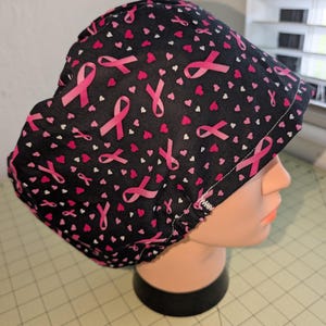 Puede incluir: Un gorro quirúrgico negro con un estampado de lazos rosas y pequeños corazones rosas y blancos. El gorro está diseñado para cubrir la cabeza y el cabello, comúnmente usado en entornos médicos.