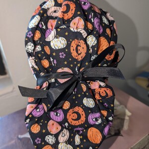 May include: A black scrub cap with a Halloween pumpkin pattern in orange, purple, and white, with gold stars and small floral details. The cap is tied with a black ribbon bow.